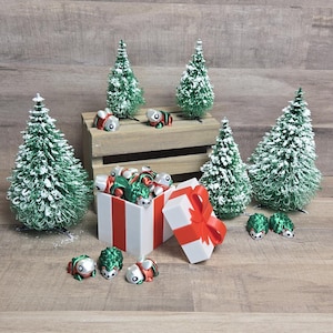 Puede incluir: Una caja de regalo roja y blanca con una tapa verde y plateada está abierta, revelando una colección de pequeñas figuras verdes y plateadas. La caja está rodeada de árboles de Navidad en miniatura cubiertos de nieve y más figuras.
