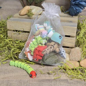 May include: A sheer white bag filled with small, colourful plush toy creatures, including dinosaurs and other whimsical designs. A green and red caterpillar toy is in the foreground. The toys are on a wooden surface. The bag is on a wooden crate.