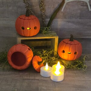 May include: Orange ceramic pumpkin decorations with cut-out eyes and mouths, lit by small LED candles. The pumpkins have dark brown stems and sit on a wooden crate with decorative moss. A festive Halloween or fall-themed display.