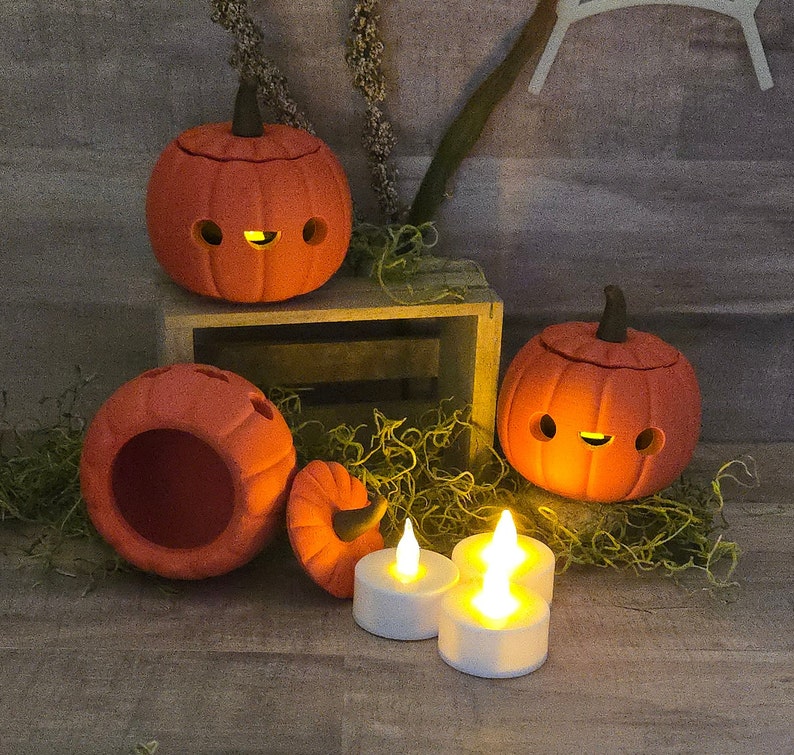 May include: Orange ceramic pumpkin decorations with cut-out eyes and mouths, lit by small LED candles. The pumpkins have dark brown stems and sit on a wooden crate with decorative moss. A festive Halloween or fall-themed display.