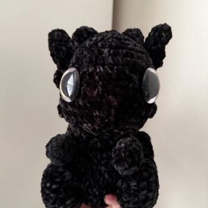 May include: Black crocheted plush toy with large eyes. The toy is shaped like a small dragon with a rounded body and a small tail.
