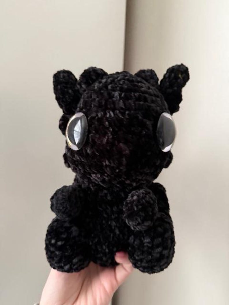 May include: Black crocheted plush toy with large eyes. The toy is shaped like a small dragon with a rounded body and a small tail.
