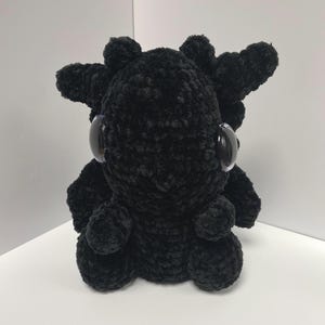 May include: A black crocheted plush toy with large, round eyes. The toy is shaped like a small creature with a rounded body and spiky, crocheted horns.