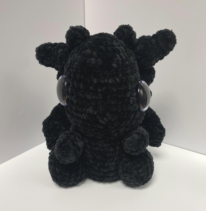 May include: A black crocheted plush toy with large, round eyes. The toy is shaped like a small creature with a rounded body and spiky, crocheted horns.