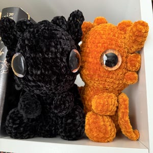 May include: Two crocheted plush toys, one black and one orange, with large googly eyes. The toys are sitting on a white shelf.