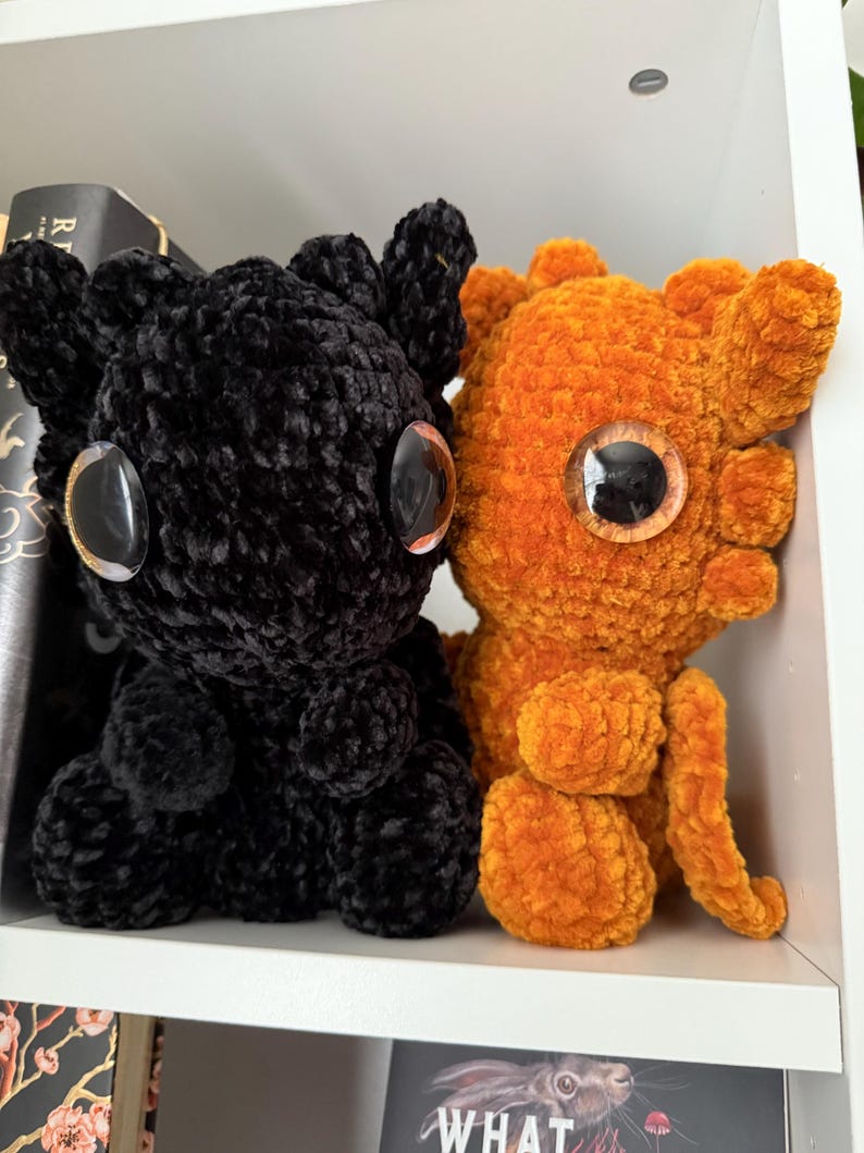 May include: Two crocheted plush toys, one black and one orange, with large googly eyes. The toys are sitting on a white shelf.