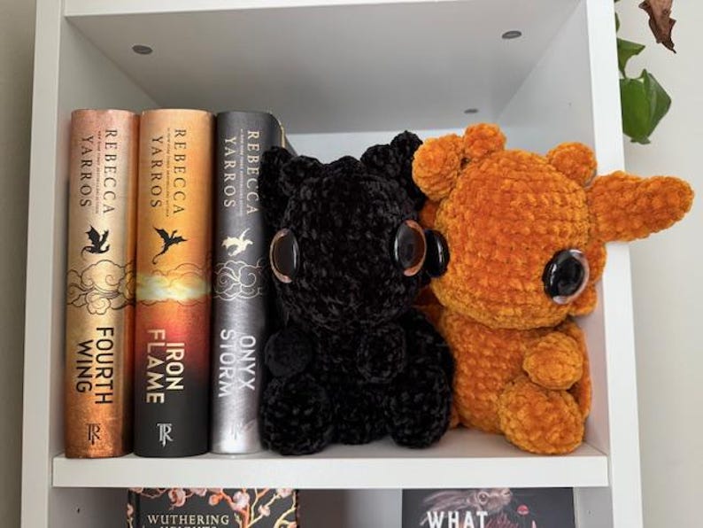 May include: Two crocheted dragon plushies on a white bookshelf. The dragon on the left is black with large black eyes. The dragon on the right is orange with large black eyes. The bookshelf is filled with books with the titles "Onyx Storm", "Iron Flame", and "Fourth Wing" visible.
