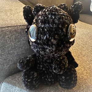 Puede incluir: Un dragón de peluche negro tejido a crochet con grandes ojos plateados. El dragón está sentado en un sofá gris.