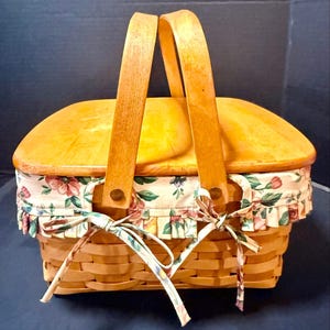 Può includere: Un cesto rettangolare in legno intrecciato con un coperchio in legno liscio color marrone chiaro e due manici verticali. Il cesto presenta una fodera in tessuto color crema con un motivo floreale rosa, verde e rosso, rifinito con bordo a volant e lacci.