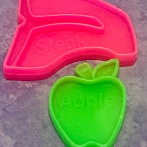 May include: A green plastic apple cookie cutter with the word "Apple" embossed on the front. The cutter is sitting on top of a pink plastic cookie cutter shaped like a half-circle.
