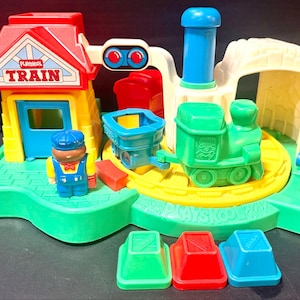 May include: A Playskool plastic train station playset featuring a yellow building with a red roof labeled TRAIN, a green locomotive, a blue cargo car, a conductor figure, a tunnel, and three separate colored blocks in green, red, and blue.