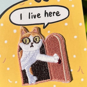 以下が含まれることがあります： オレンジ色の斑点のある黄色と白の猫が、猫の上に「ここに住んでいます」と書かれた吹き出しのある茶色のドアから顔をのぞかせています。