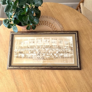 May include: A framed black and white photograph of a large group of people standing in front of a building. The photo is in a brown frame with a gold trim.