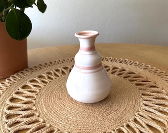 May include: A small, white ceramic vase with a rounded body and a narrow neck. The vase features two horizontal bands of pink around the body and neck. It sits on a round, woven jute placemat.