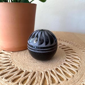 May include: A spherical black stone object featuring a carved pattern of light gray petal-like shapes around the top opening and two horizontal bands encircling the middle section, creating a contrast between the dark polished stone and the gray carvings.