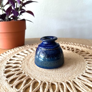 May include: A small, round ceramic vessel with a glossy, deep blue glaze. The object features a wide, rounded base that tapers upward to a narrow, circular opening with a distinct, raised rim. The surface shows subtle variations in blue tones.