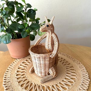 May include: A woven kangaroo-shaped basket with a round basket for a pouch. The kangaroo is made of light brown wicker and has black eyes. It sits on a round, woven placemat. A potted plant is in the background.