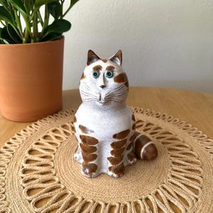 May include: A ceramic cat figurine featuring a white base with brown tabby stripes, pointed ears, and distinct teal-colored eyes. The cat is depicted in a seated position with its tail curled to the side, showcasing a smooth, glazed finish.