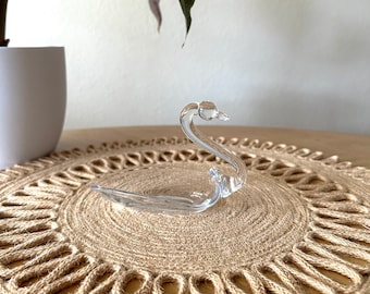 May include: A clear glass swan figurine on a brown woven placemat.