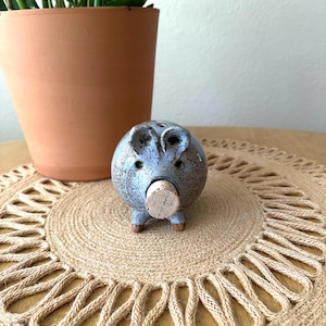 May include: A small, round, ceramic pig figurine with a speckled light blue glaze. The pig features two upright ears, two small eyes, and a light-colored cylindrical cork plug inserted into the snout area. It has four small, brown, unglazed feet.