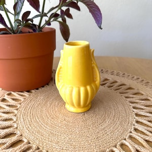 May include: A small, bright yellow ceramic vase with a ribbed design and two decorative handles. The vase is displayed on a round, woven jute placemat with a fringed edge. A potted plant with purple leaves is in the background.