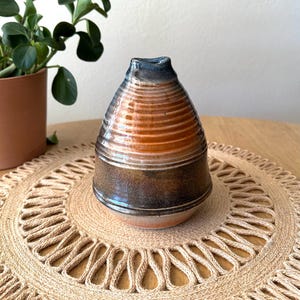 May include: A handmade ceramic vase with a unique shape and color gradient. The vase transitions from a dark brown base to a light orange middle, with a dark blue rim. The vase is sitting on a round, woven placemat.