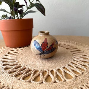 May include: A small, round, beige ceramic vase with a painted butterfly design in red, black, and blue. The vase sits on a round, woven jute placemat. A potted plant with purple and green leaves is in the background.