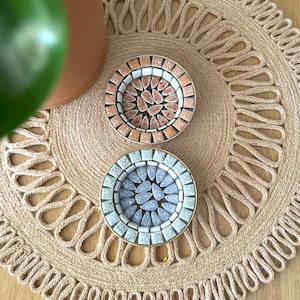 May include: Two circular mosaic coasters featuring intricate patterns. The top coaster displays warm peach and terracotta tones, while the bottom coaster features cool blue and grey shades. Both have a metallic rim and a central petal design.
