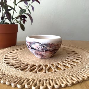 May include: A small, round ceramic bowl with a marbled design in shades of pink, white, and black. The bowl sits on a round, woven jute placemat. A potted plant with purple leaves is in the background.