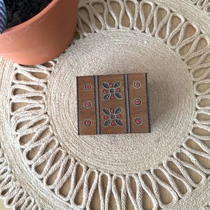 May include: A small, rectangular wooden box with a decorative floral design in red and black. The box is brown with red circles and is placed on a beige woven placemat. A terracotta pot with a plant is in the background.