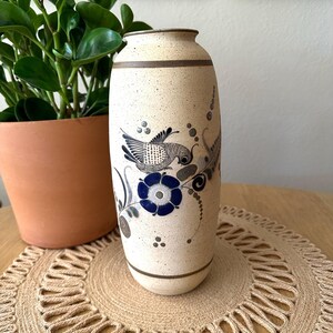 May include: A cylindrical ceramic vase with a speckled cream-colored finish, featuring hand-painted dark blue floral motifs and a detailed black bird illustration. The vase is accented with horizontal brown bands at the top and bottom edges.