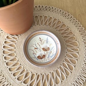May include: A small, round ceramic dish featuring a speckled cream-colored base with a hand-painted botanical design in shades of brown and blue. The dish has a raised rim with a brown band and sits centered on a circular, woven tan jute placemat.