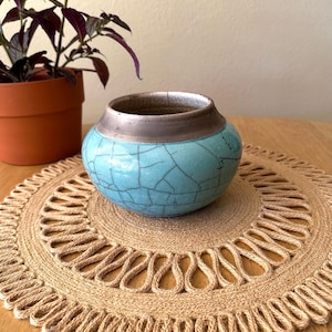 May include: A turquoise ceramic bowl with a crackle glaze and a silver rim sits on a round, woven jute placemat. A potted plant with purple leaves is in the background. The bowl is a decorative item.