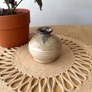 May include: A small, round ceramic vase with a beige and brown color scheme. The vase has a narrow neck and a textured surface. It sits on a woven jute placemat. A potted plant is visible in the background.
