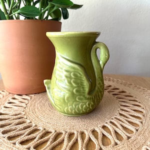 May include: A small, light green ceramic pitcher shaped like a swan. The body of the pitcher features embossed feather details, while the handle is formed by the swan's long, curved neck. The piece has a smooth, glossy finish and a wide, flared rim.