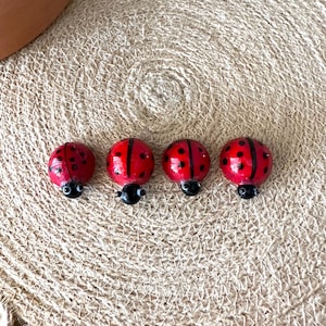 May include: Four small, rounded ladybug figurines arranged in a horizontal row. Each ladybug features a bright red back with distinct black spots, a glossy finish, and a small black head with two tiny white eyes protruding from the front.
