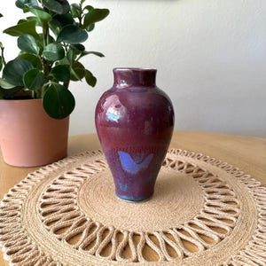 May include: A handcrafted ceramic vase with a glossy, deep burgundy glaze. The vase has a rounded body and a slightly flared rim. It sits on a round, woven jute placemat. A potted plant is visible in the background.