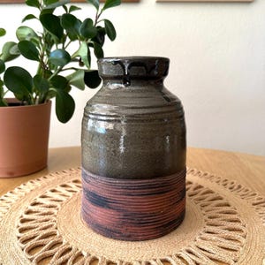 May include: A handcrafted ceramic vase with a dark brown top and a textured, earthy green body. The base features horizontal bands of reddish-brown. The vase is displayed on a round, woven placemat.