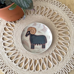 May include: A small, round ceramic plate with a black ram illustration, featuring a brown curly horn design. The plate is set on a beige woven placemat with a decorative edge. A potted plant is visible in the background.
