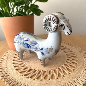 May include: A white ceramic ram figurine featuring hand-painted blue and brown floral patterns across its body. The ram has curved, striped horns and detailed line work on its face, standing on four short legs with brown striped accents on the feet.