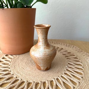 May include: A small ceramic bud vase with a flared rim and a bulbous body. The vase features a speckled, mottled glaze in shades of tan, cream, and light brown, with a smooth, glossy finish that highlights the natural variations in the clay.