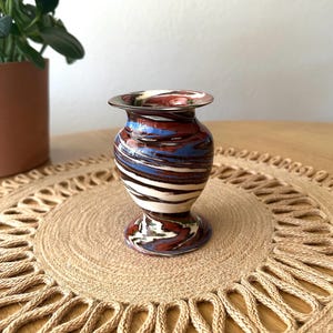 May include: A small, decorative vase with a pedestal base and flared rim. The vase features a swirled pattern of red, blue, white, and brown. The vase is approximately 10 cm tall and sits on a round, woven placemat.