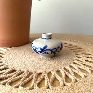 May include: A small, squat ceramic vase with a cream-colored glaze and a hand-painted blue swirling vine pattern encircling the body. The vessel features a narrow, short neck and a wide, rounded base, showcasing a smooth, glossy finish.