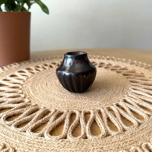 May include: A small, dark brown ceramic vase with a glossy finish. The vase has a ribbed texture and a rounded shape, sitting on a natural fiber placemat. A potted plant is visible in the background.