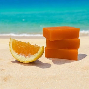 May include: Three orange bars of soap stacked on top of each other on a sandy beach. A slice of orange is in the foreground.