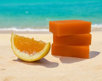 May include: Three orange bars of soap stacked on top of each other on a sandy beach. A slice of orange is in the foreground.