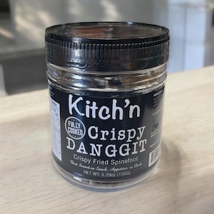 Puede incluir: Un tarro de vidrio transparente con tapa negra que contiene danggit frito crujiente, un plato de pescado seco filipino. El tarro tiene una etiqueta negra con texto blanco que dice "Kitch'n Fully Cooked Crispy DANGGIT Crispy Fried Spinefoot Best Served as Snack, Appetizer or Dish NET WT: 5.29oz (150G)"