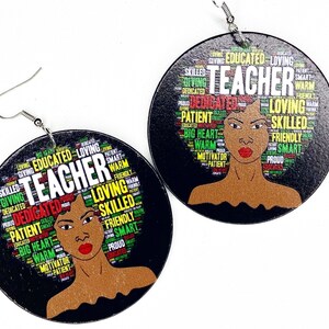May include: A pair of wooden earrings with a black background and a colorful word cloud depicting the qualities of a teacher. The word cloud is shaped like an afro hairstyle and features a woman's face in the center. The earrings are a great way to show your appreciation for teachers.