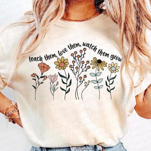 May include: A white t-shirt with the text "teach them, love them, watch them grow" printed on it. The text is surrounded by a row of colorful flowers.