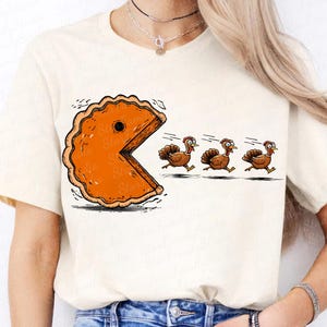 May include: A cream-colored short-sleeve t-shirt featuring a graphic print of a large orange pumpkin pie slice shaped like a video game character chasing three small brown turkeys across the front of the garment.